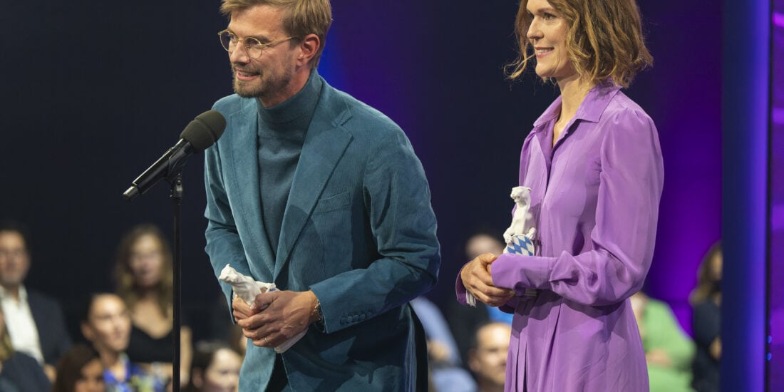 Executive Producer Julia Mehnert (rechts) und Moderator Joko Winterscheidt (links) bei der Preisverleihung. Joko steht am Mikrofon und hält eine Dankesrede.