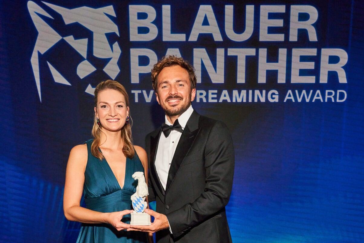 Judith Gerlach und David Helmut bei der Preisverleihung des Blauen Panthers 2022. Beide haben die Hand an der Panther Statue.