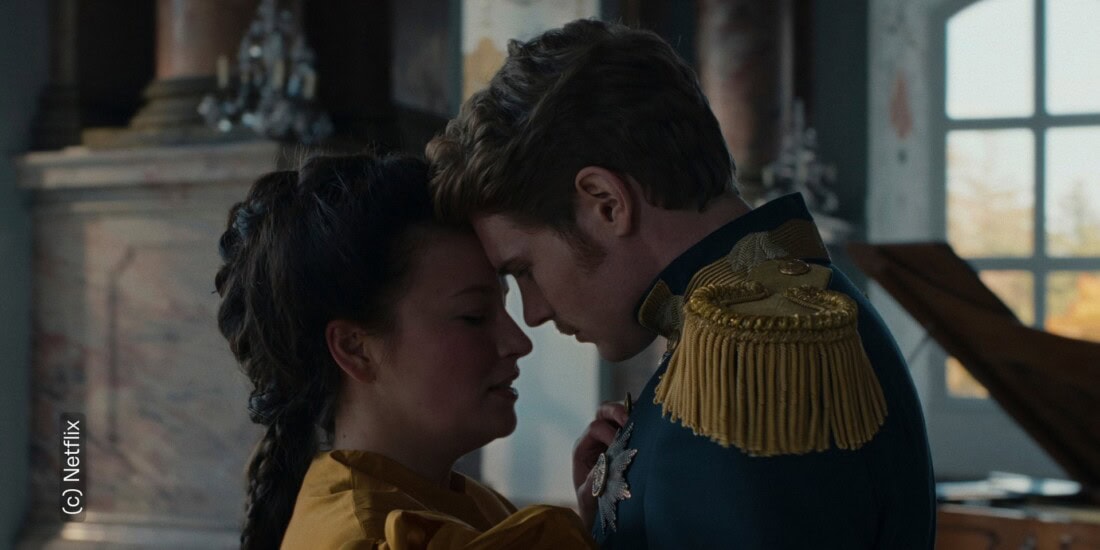 Zu sehen sind Sisi und Kaiser Franz. Sie stehen nah beieinander. Ihre Stirne lehnen aneinander. Ihre Gesichtsausdrücke wirken verzweifelt, als wären sie innerlich zerrissen.