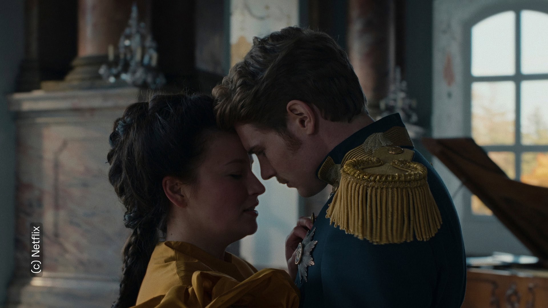 Zu sehen sind Sisi und Kaiser Franz. Sie stehen nah beieinander. Ihre Stirne lehnen aneinander. Ihre Gesichtsausdrücke wirken verzweifelt, als wären sie innerlich zerrissen.