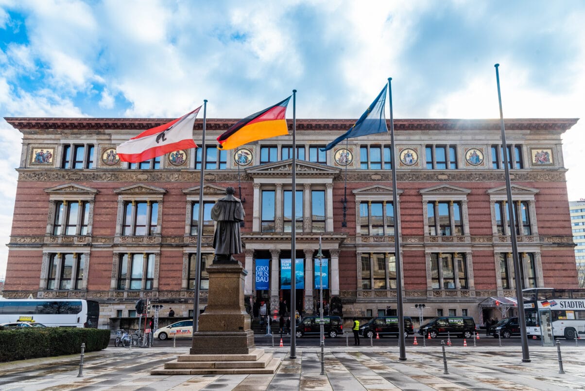 Ein großes altes schönes Gebäude. Am Eingang hängen Werbefahnen des Gropius Baus. Davor zu sehen in eine Statue, die dem Betrachter dem Rücken zukehrt. Außerdem mehrere Flaggenmäste mit der Berliner Flagge, der Deutschlandflagge und der Europa Flagge.
