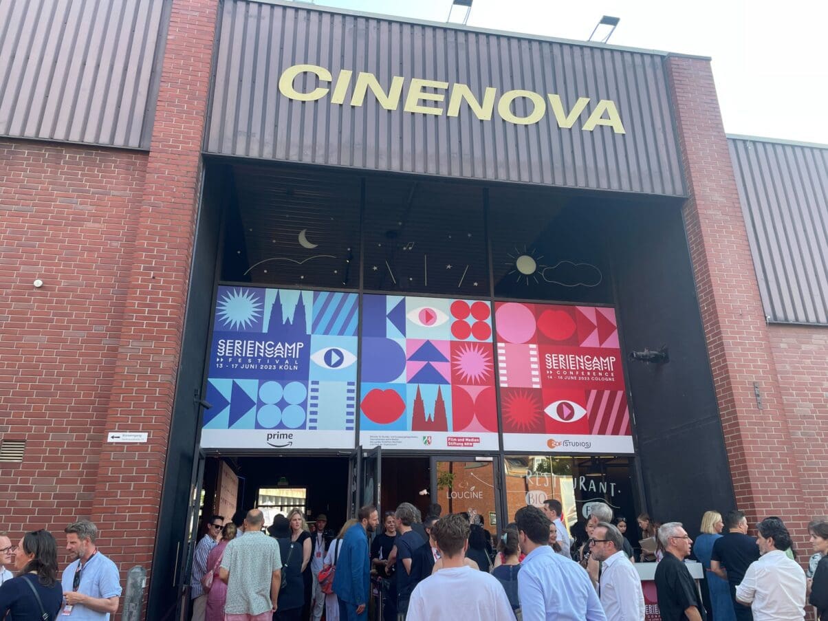 Das Cinenova Kino in Köln Ehrenfeld von Außen. Der Eingang ist mit dem blau-roten Design des Seriencamp Festivals & Seriencamp Conference gebranded. Vor dem Eingang stehen einige Leute und sprechen miteinander.