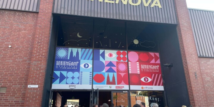 Das Cinenova Kino in Köln Ehrenfeld von Außen. Der Eingang ist mit dem blau-roten Design des Seriencamp Festivals & Seriencamp Conference gebranded. Vor dem Eingang stehen einige Leute und sprechen miteinander.