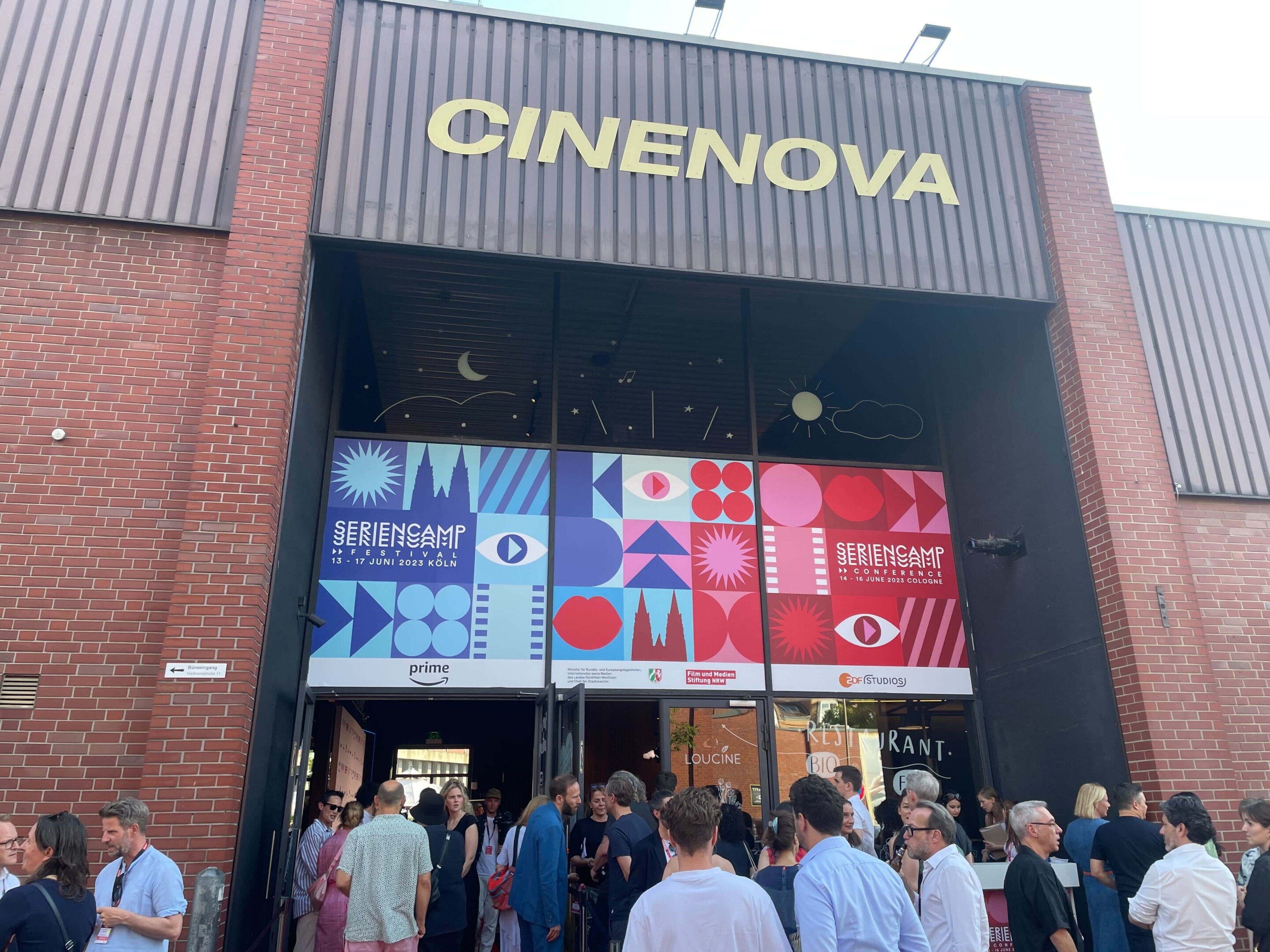 Das Cinenova Kino in Köln Ehrenfeld von Außen. Der Eingang ist mit dem blau-roten Design des Seriencamp Festivals & Seriencamp Conference gebranded. Vor dem Eingang stehen einige Leute und sprechen miteinander.