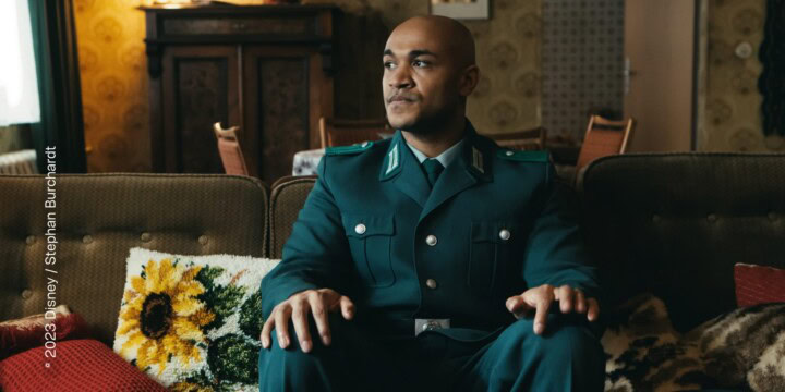 Ein Mann sitzt in einer dunkelgrünen Uniform auf einem Sofa in einem gemütlichen Wohnzimmer. Er sitzt in der Mitte des Sofas, sein Kopf geht in Richtung des Fensters. Sein Blick ist konzentriert und besorgt. Der Mann ist PoC.