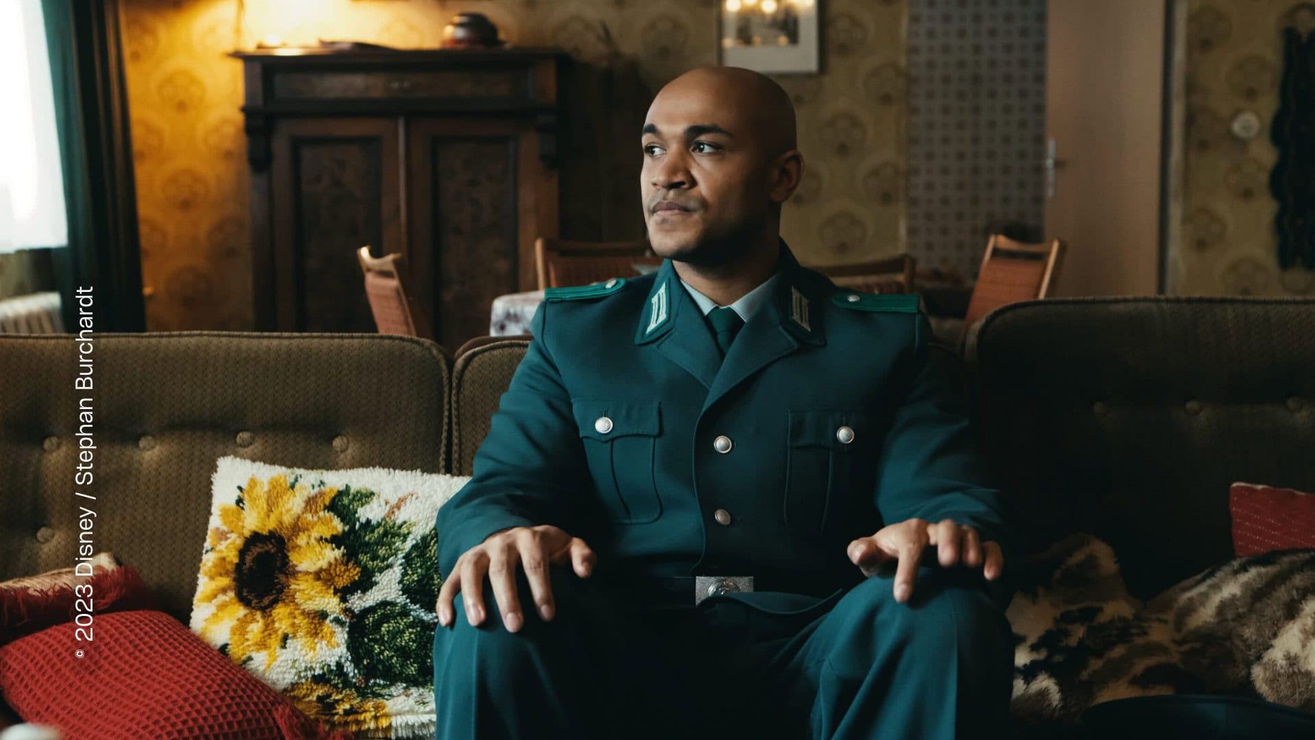 Ein Mann sitzt in einer dunkelgrünen Uniform auf einem Sofa in einem gemütlichen Wohnzimmer. Er sitzt in der Mitte des Sofas, sein Kopf geht in Richtung des Fensters. Sein Blick ist konzentriert und besorgt. Der Mann ist PoC.