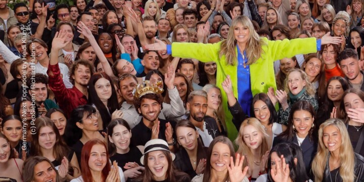 Heidi Klum steht mit ausgebreiteten Armen in einer Menschenmenge, bestehend aus jungen Erwachsenen. Alle lächeln in die Kamera, viele jubelnd und klatschen. Heidi Klum steht leicht erhöht und trägt ienen bunten Mantel. Aus: Germany's Next Topmodel by Heidi Klum, Staffel 19. (c) ProSieben / Richard Hübner.