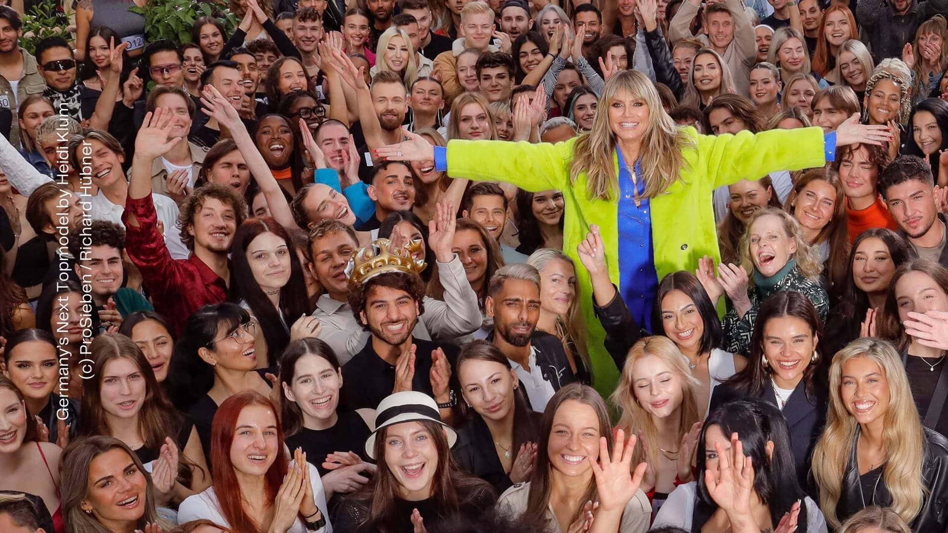 Heidi Klum steht mit ausgebreiteten Armen in einer Menschenmenge, bestehend aus jungen Erwachsenen. Alle lächeln in die Kamera, viele jubelnd und klatschen. Heidi Klum steht leicht erhöht und trägt ienen bunten Mantel. Aus: Germany's Next Topmodel by Heidi Klum, Staffel 19. (c) ProSieben / Richard Hübner.