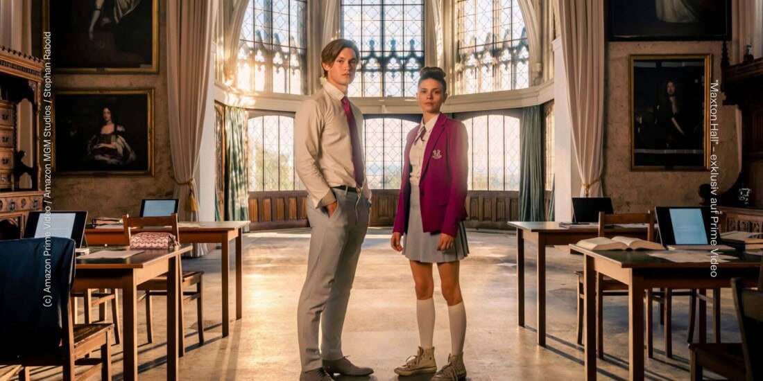 Die Protagonist:innen der Serie von "Maxton Hall - Die Welt zwischen uns" stehen in einem großen Lehrsaal, der sich in einer Art mittelalterlichen Burg befindet. Beide Personen tragen eine Schuluniform. Um sie herum stehen Tische, auf denen sich Lernutensilien wie Tablets, Bücher, Papier und Mäppchen stehen. An den Wänden hängen alte Bilder.