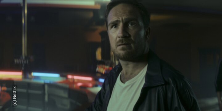 Protagonist (gespielt von Frederik Lau) der Serie "Crooks" von Netflix sieht erstaunt und knapp an der Kamera vorbei. Er befindet sich in einem dunklen Raum. Im Hintergrund sieht verschwommen ein paar Lichter in verschiedenen Farben zu erahnen.