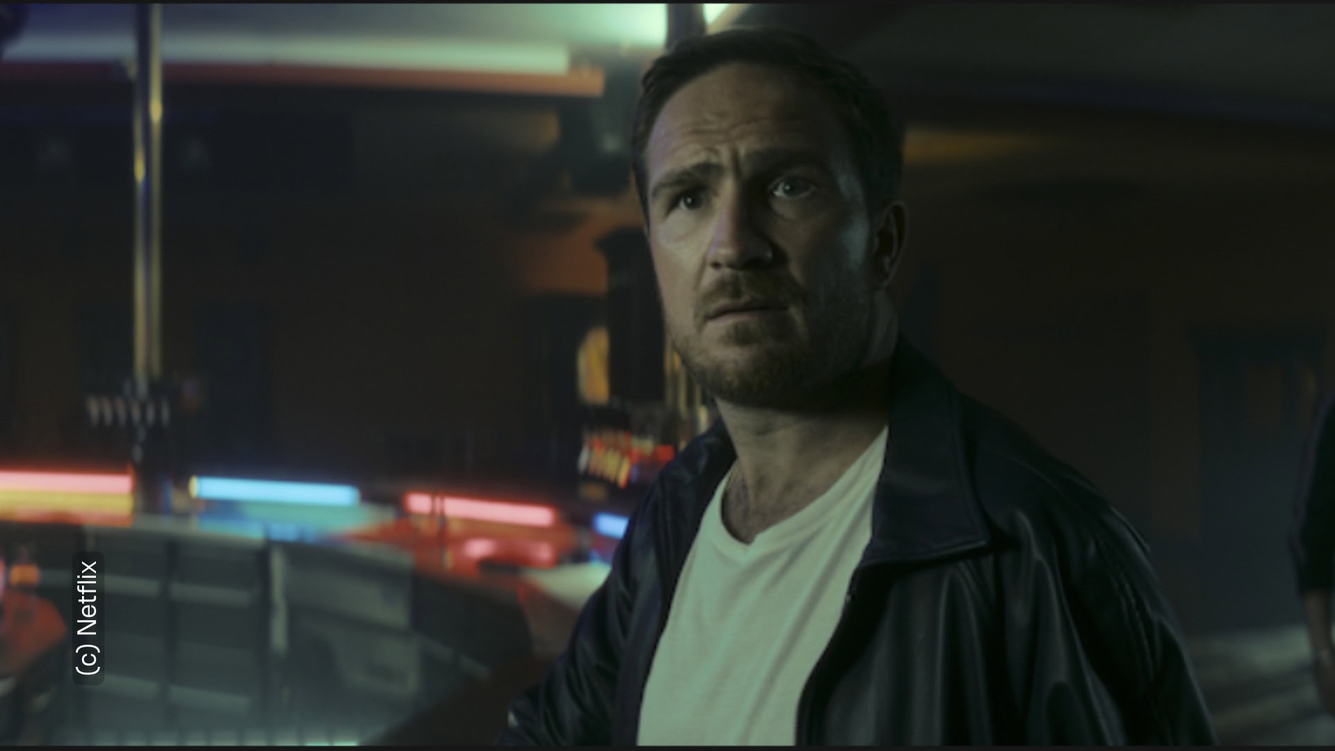 Protagonist (gespielt von Frederik Lau) der Serie "Crooks" von Netflix sieht erstaunt und knapp an der Kamera vorbei. Er befindet sich in einem dunklen Raum. Im Hintergrund sieht verschwommen ein paar Lichter in verschiedenen Farben zu erahnen.