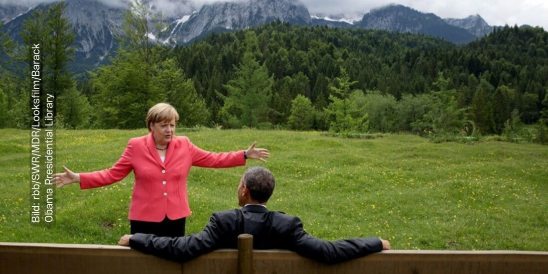 Angela Merkel steht mit ausgebreiteten Armen vor dem ehemaligen US-Präsidenten Barack Obama. Er sitzt auf einer Holzbank, sein Rücken ist zur Kamera gedreht. Obama sitzt entspannt, seine Arme liegen ausgebreitet auf dem Rücken der Holzbank. Angela Merkel scheint ihm etwas zu erklären. Hinter Angela Merkel ist eine naturbelassene Landschaft zu sehen: Eine grüne Wiese, dahinter ein dunkelgrüner Wald und dahinter eine wunderschöne Berglandschaft.