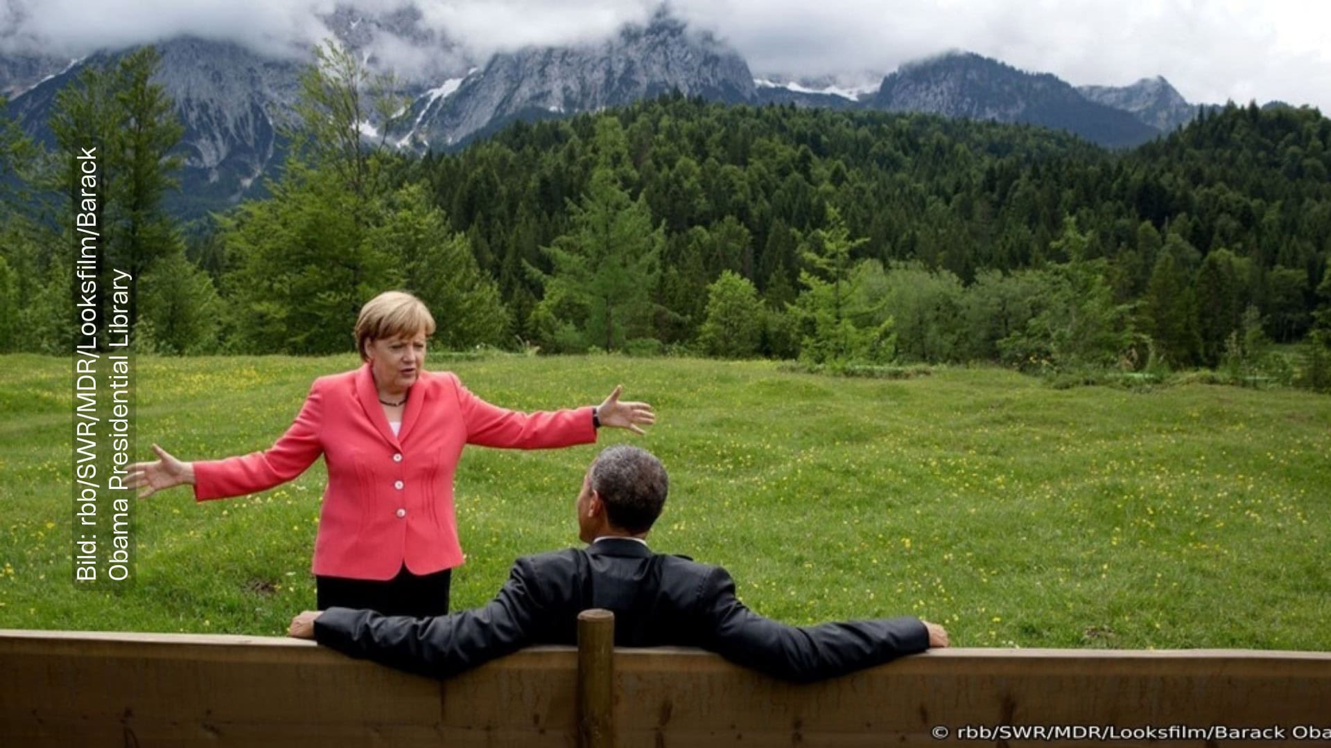 Angela Merkel steht mit ausgebreiteten Armen vor dem ehemaligen US-Präsidenten Barack Obama. Er sitzt auf einer Holzbank, sein Rücken ist zur Kamera gedreht. Obama sitzt entspannt, seine Arme liegen ausgebreitet auf dem Rücken der Holzbank. Angela Merkel scheint ihm etwas zu erklären. Hinter Angela Merkel ist eine naturbelassene Landschaft zu sehen: Eine grüne Wiese, dahinter ein dunkelgrüner Wald und dahinter eine wunderschöne Berglandschaft.