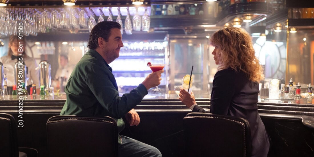 Die Hauptfiguren, gespielt von Bastian Pastewka und Anke Engelke sitzen an einer Bar-Theke. Ihre Körper sind zueinander gerichtet, sie sind dabei mit ihren Cocktails anzustoßen. „Perfekt verpasst" von Prime Video (c) Amazon MGM Studios / Ben Knabe