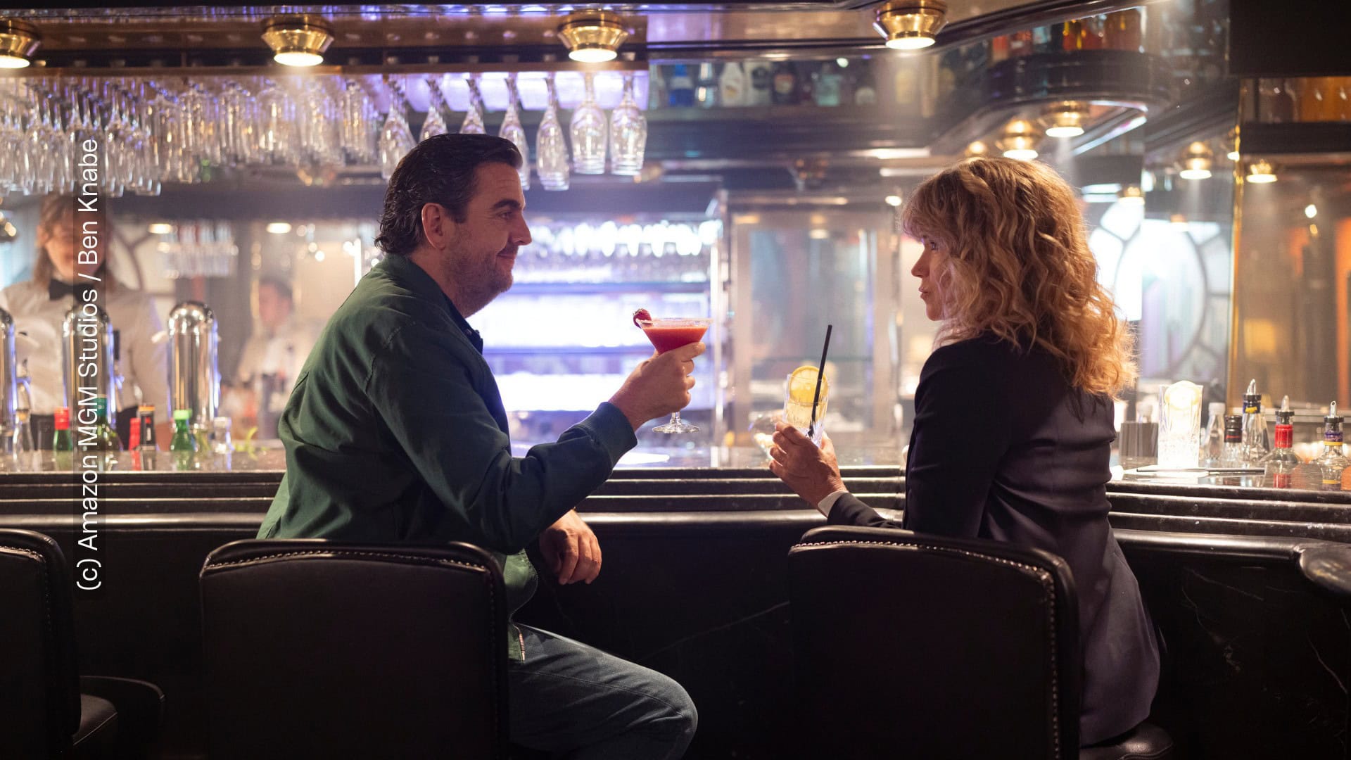 Die Hauptfiguren, gespielt von Bastian Pastewka und Anke Engelke sitzen an einer Bar-Theke. Ihre Körper sind zueinander gerichtet, sie sind dabei mit ihren Cocktails anzustoßen. „Perfekt verpasst" von Prime Video (c) Amazon MGM Studios / Ben Knabe