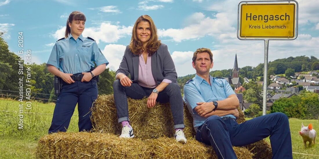 Aus: Mord mit Aussicht – Staffel 5. Die Kommissarin sitzt zusammen mit einem in Uniform gekleideten Polizisten auf einem Stroh-Ballen. Daneben steht eine weitere Polizistin in Uniform. Die Kommissarin trägt einen Blazer und legere Kleidung - Bluse, Jeans, Sneaker. Sie blicken in die Kamera. Daneben ist ein Orsschild mit dem Text "Hengasch - Kreis Liebernich" zu sehen. Im Hintergrund sind viele Häuser und eine Kirche aus einem Dorf zu sehen. Schräg hinter den drei Personen ist ein kleines Schwein.