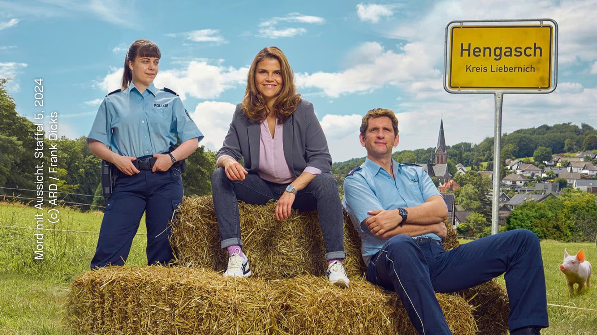 Aus: Mord mit Aussicht – Staffel 5. Die Kommissarin sitzt zusammen mit einem in Uniform gekleideten Polizisten auf einem Stroh-Ballen. Daneben steht eine weitere Polizistin in Uniform. Die Kommissarin trägt einen Blazer und legere Kleidung - Bluse, Jeans, Sneaker. Sie blicken in die Kamera. Daneben ist ein Orsschild mit dem Text "Hengasch - Kreis Liebernich" zu sehen. Im Hintergrund sind viele Häuser und eine Kirche aus einem Dorf zu sehen. Schräg hinter den drei Personen ist ein kleines Schwein.
