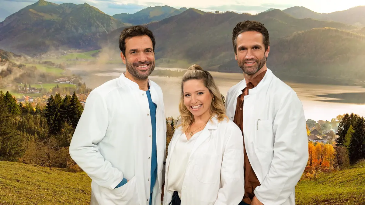 Die Hauptfiguren stehen in der Mittel. EIne Frau und zwei Männer. Alle tragen Arztkittel. Im Hintergrund ist eine wunderschöne Landschaft im Sonnenlicht zu sehen - mit Bergen, einem See und einer grünen Wiese.