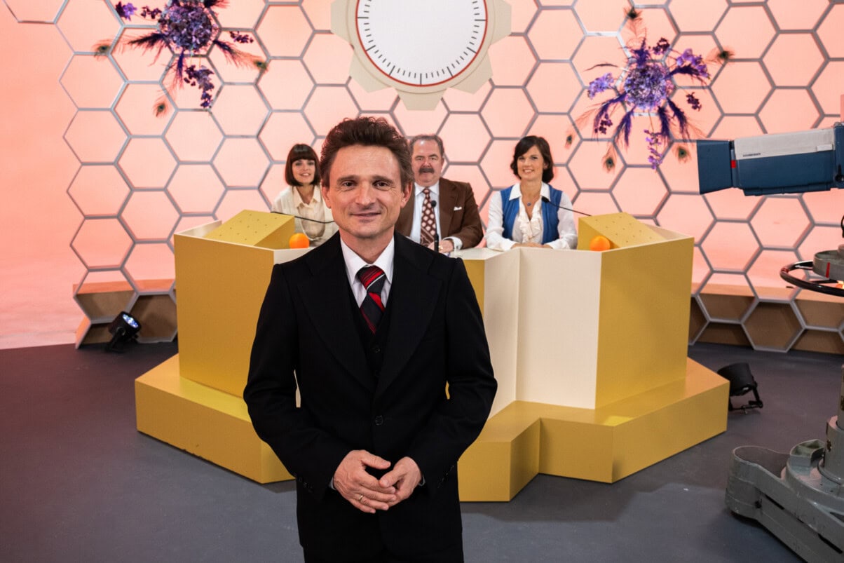 TV & Streaming Tipps Oktober: Moderator in einem Fernsehstudio, Teilnehmer:innen an einem Pult im Hintergrund