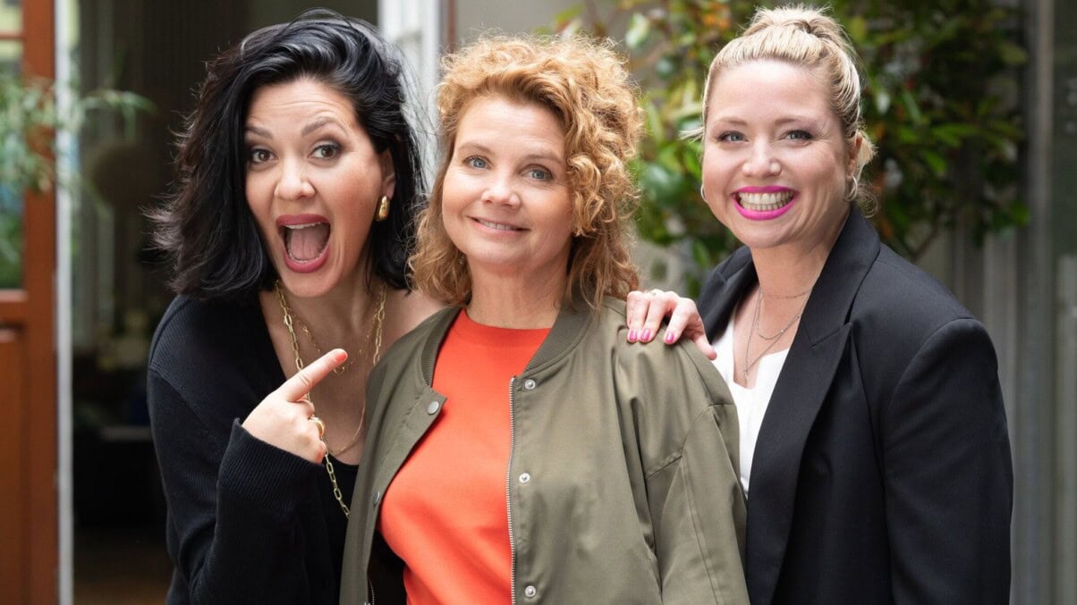 TV & Streaming Tipps November: Drei Frauen stehen lachend nebeneinander und schauen in die Kamera; eine von ihnen zeigt mit einem Finger auf die Frau in der Mitte, alle wirken fröhlich und energiegeladen.