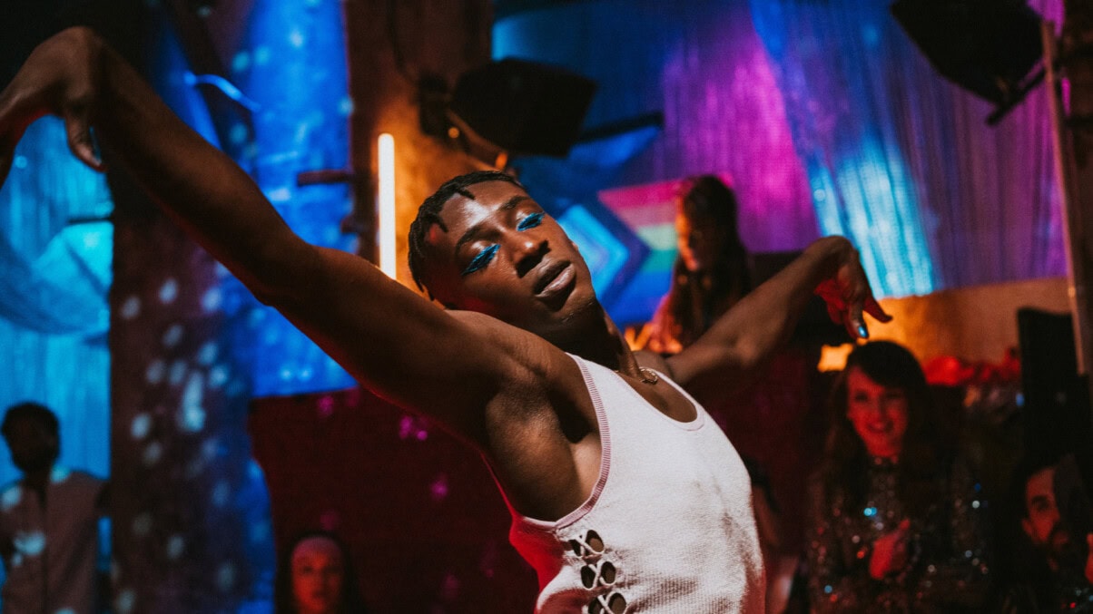TV & Streaming Tipps Dezember 2025: Eine Person mit blauem Augen-Make-up tanzt in einem Club mit weit ausgebreiteten Armen, trägt ein weißes Top und ist von buntem Neonlicht sowie unscharfen Personen im Hintergrund umgeben.