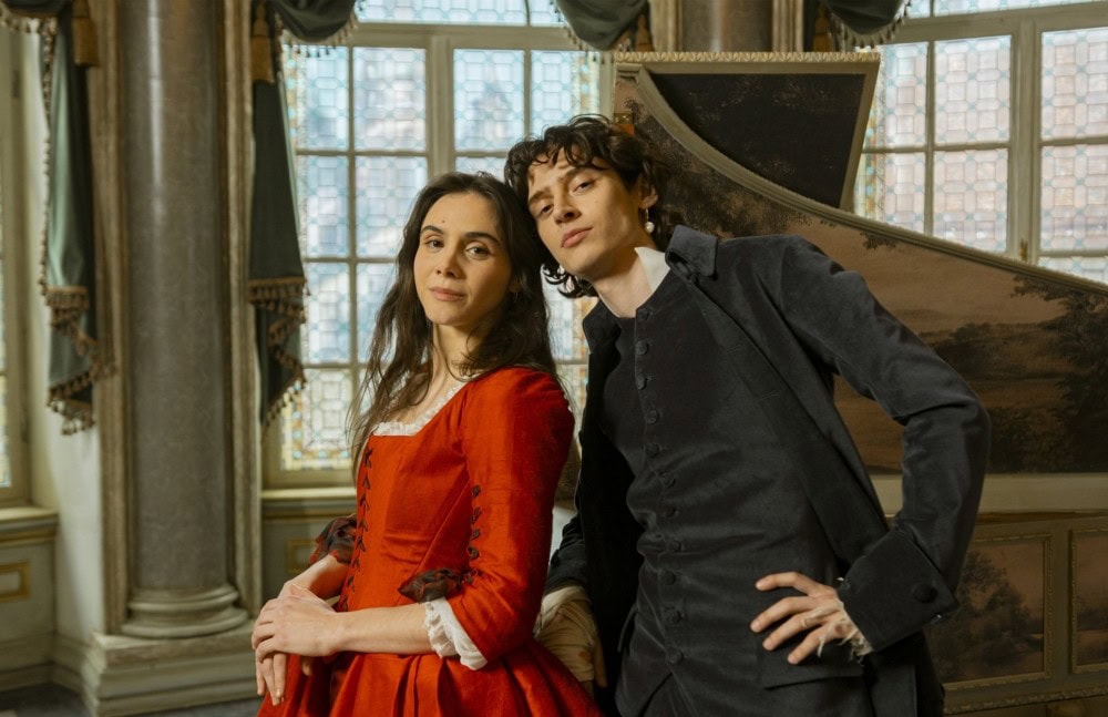 TV & Streaming Tipps Dezember 2025: Zwei junge Erwachsene in historischer Kleidung – sie im roten Kleid, er im dunklen Anzug – lehnen selbstbewusst an einem Cembalo in einem prunkvollen Raum mit großen Fenstern.