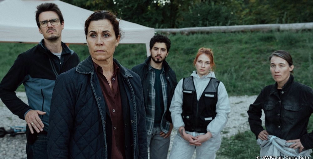 Fünf Personen stehen in einer grünen Landschaft. Zwei der drei Frrauen tragen eine schusssichere Weste. Die Frau, die keine Weste trägt, steht im Vordergrund. Alle Personen schauen knapp an der Kamera vorbei. Die Stimmung wirkt gedrückt. Es ist Dämmerung.