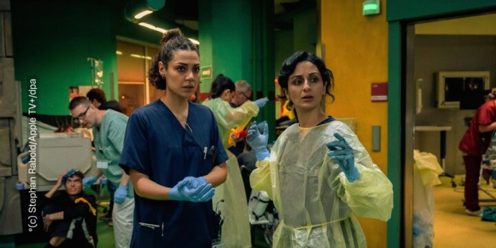 Zwei Ärzt:innen stehen in einem Saal. Sie schauen an der Kamera vorbei. Um sie herum stehen weitere Krankenhausangestellte, die beschäftigt sind, Kranke und Verwundete zu versorgen. Die Farben sind dunkel gehalten, die Atmosphäre wirkt hektisch.