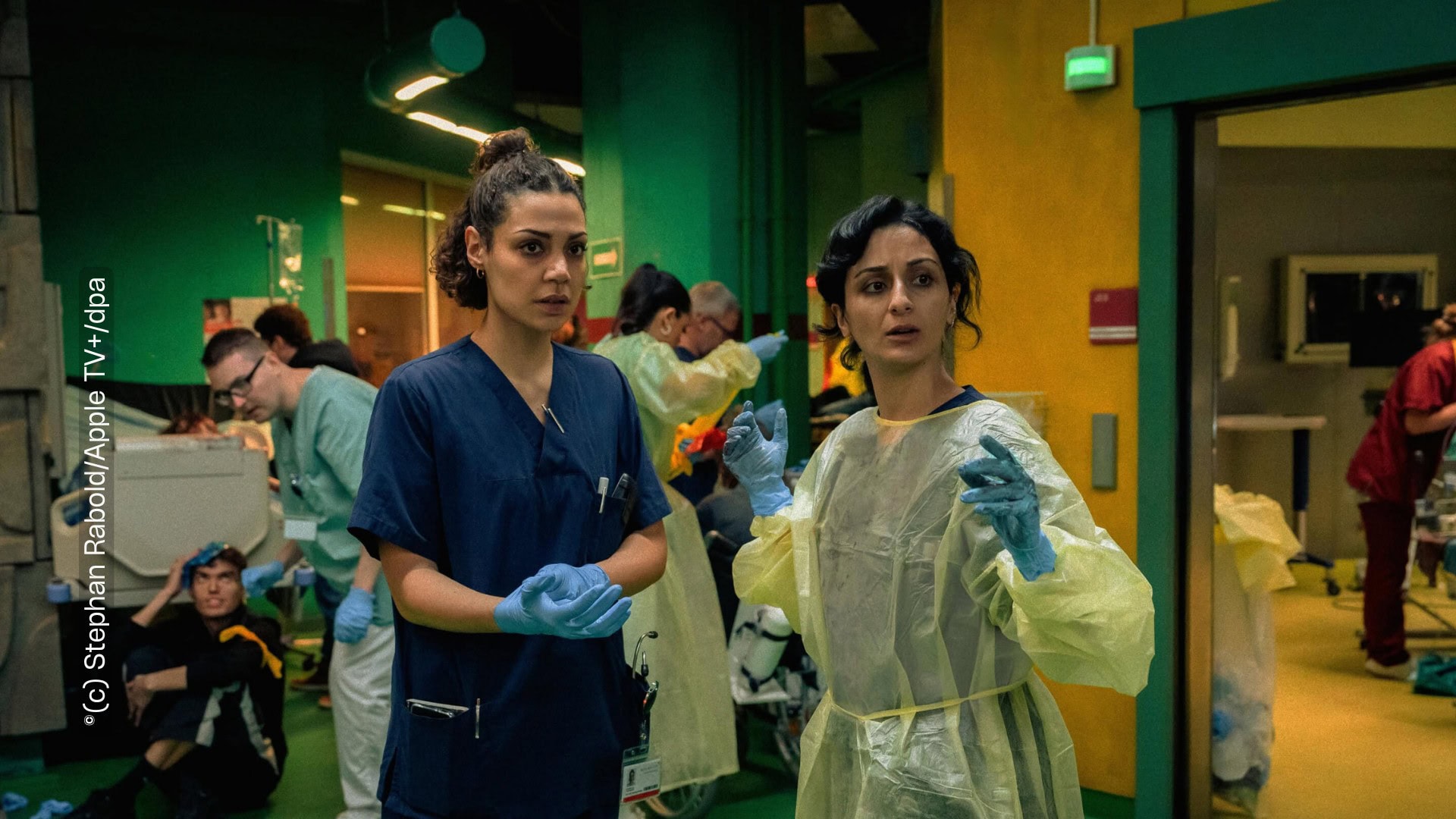 Zwei Ärzt:innen stehen in einem Saal. Sie schauen an der Kamera vorbei. Um sie herum stehen weitere Krankenhausangestellte, die beschäftigt sind, Kranke und Verwundete zu versorgen. Die Farben sind dunkel gehalten, die Atmosphäre wirkt hektisch.