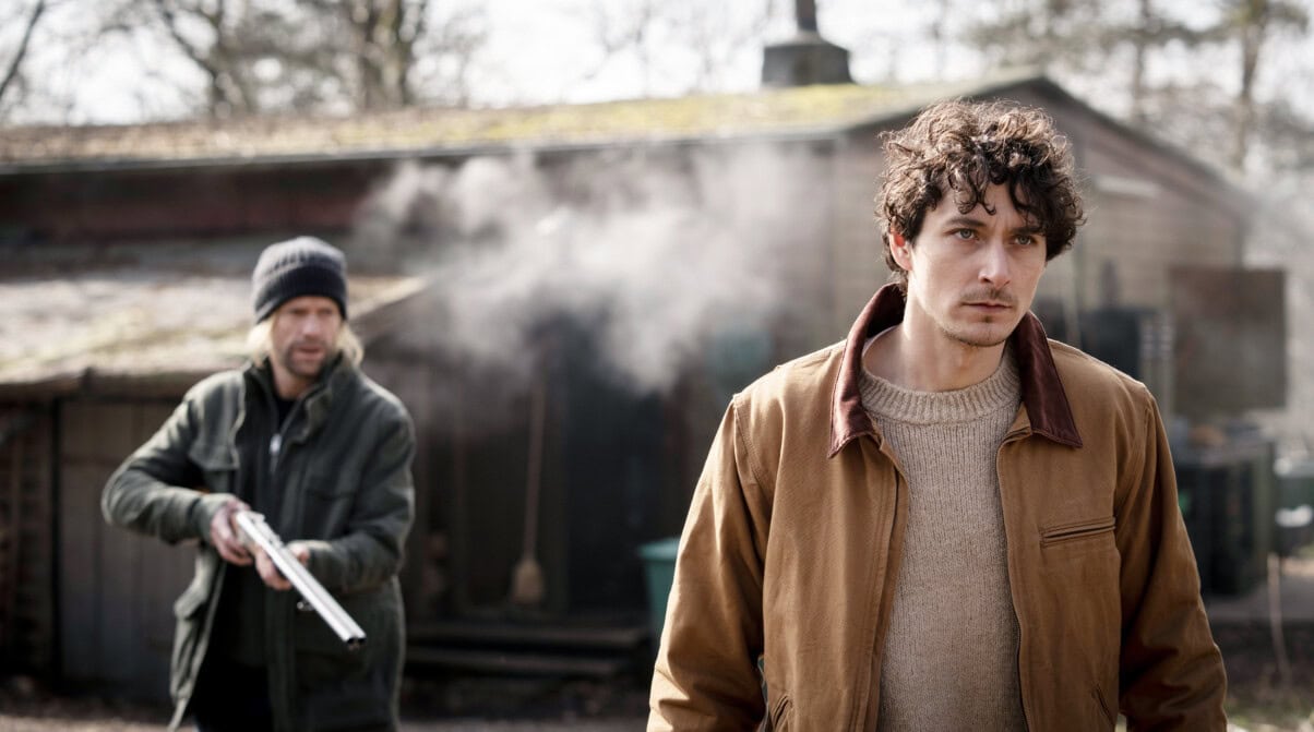 Zwei Männer stehen in einer trüben naturbelassenen Landschaft. Hinter ihnen steht ein Holzhaus. Der Mann rechts schaut bestimmt in die Ferne. Hinter ihm steht ein Mann mit einer Mütze mit einem Gewehr. Sein Blick ist offen, es wirkt, als hätte er gare einen Schuss losgelassen, da zwischen den beiden Männern eine Rauchwolke zu erkennen ist. Das Gewehr ist leicht gesenkt, die Stimmung wirkt angespannt.