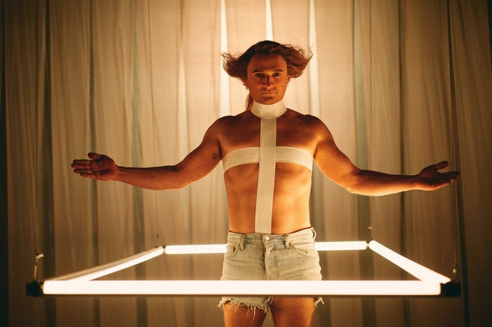 Ein Mann steht in einem viereckigen Rahmen, bestehend aus LED-Stäben. Der Mann trägt eine kurze Jeans-Short und kein Oberteil. Stattdessen hat er ein Kreuz über seine Brust mit weißem Klebeband geklebt sowie eine Art Rollkragen. Seine Arme sind ausgebreitet. Sein Blick ist starr nach vorne gerichtet und seine Haare sind an den Seiten zauselig nach oben gestylt.