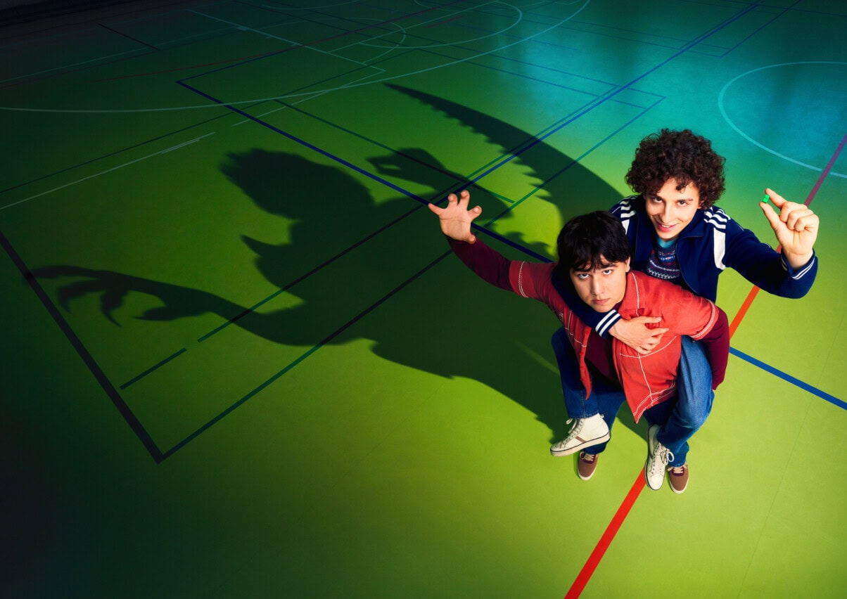 "Club der Dinosaurier": Keyvisual mit Janni (Diyar Ilhan) und Ben (Shadi Eck). Die beiden Jungen stehen in einer Turnhalle Huckepack und im Schatten sieht man Umrisse von wilden Kreaturen.