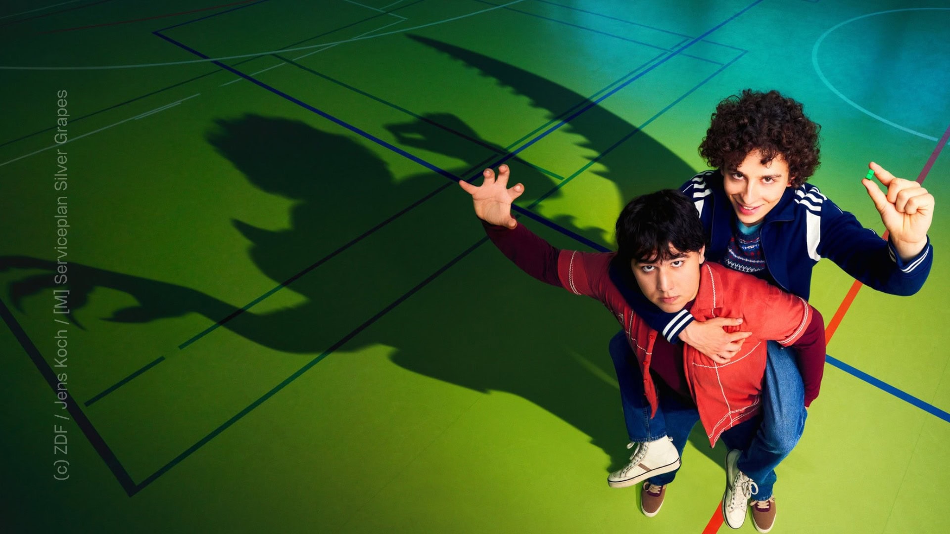 TV & Streaming Tipps im Juni: "Club der Dinosaurier": Keyvisual mit Janni (Diyar Ilhan) und Ben (Shadi Eck). Die beiden Jungen stehen in einer Turnhalle Huckepack und im Schatten sieht man Umrisse von wilden Kreaturen.