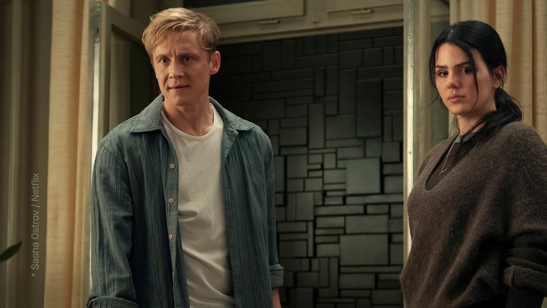 TV & Streaming Tipps im Juli 2025: Ein Mann, gespielt von Matthias Schweighöfer, und eine Frau, gespielt von Ruby O. Fee, stehen in einem Raum. Man sieht Vorhänge an einem Fenster. Das Fenster ist geöffnet. Vor dem Fenster ist keine Landschaft zu sehen, sondern eine schwarze Mauer, gebaut aus unterschiedlich geformten Mauersteinen. Offizielles Pressefoto von „Brick– Kein Weg nach draußen” (Netflix).