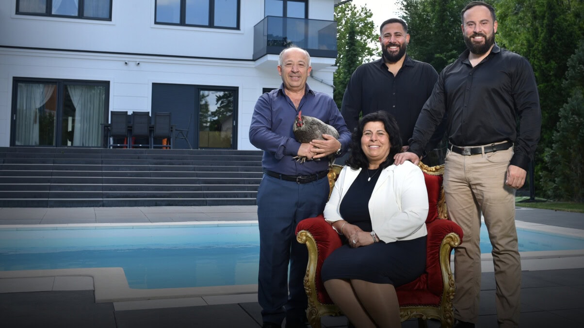 TV & Streaming Tipps im August 2025: Vier Personen stehen etwas rechts im Zentrum des Bildes, alle lächeln in die Kamera. Zwei Männer mittleren Alters in schwarzen Hemd und hellbrauner Hose, eine ältere Frau in schwarzem Etuikleid und weißem Blazer die in einem eleganten Sessel sitzt und ein älterer Mann, blau gekleidet, der ein Huhn in seinen Händen hält. IM Hintergrund ist ein großes Haus sowie ein Pool zu sehen.