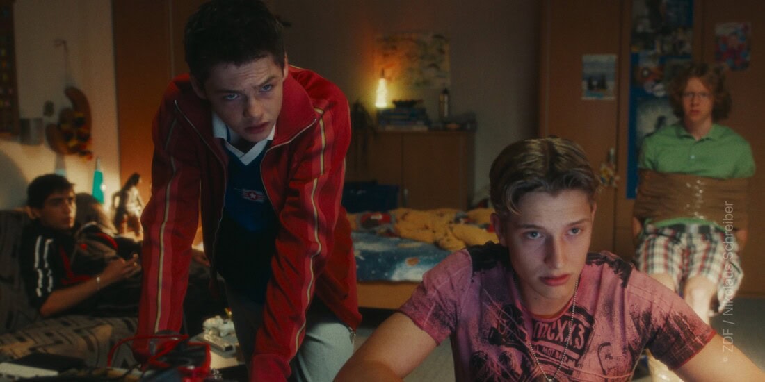 TV & Streaming Tipps im August 2025: In einem Kinderzimmer befinden sich vier Jungs im Teenager-Alter. Im Vordergrund sitzt einer am Schreibtisch an einer Tastatur, der zweite Lehnt über dem Schreibtisch und sieht in dieselbe Richtung wie der erste - vermutlich blicken beide in einen Computerbildschirm. Im Hintergrund sitzen zwei weitere Jungen - einer ist mit Paketklebeband gefesselt auf einem Stuhl, der zweite sitzt auf einer Couch - auch sie blicken beide Richtung Bildschirm.