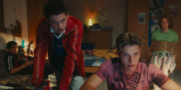 TV & Streaming Tipps im August 2025: In einem Kinderzimmer befinden sich vier Jungs im Teenager-Alter. Im Vordergrund sitzt einer am Schreibtisch an einer Tastatur, der zweite Lehnt über dem Schreibtisch und sieht in dieselbe Richtung wie der erste - vermutlich blicken beide in einen Computerbildschirm. Im Hintergrund sitzen zwei weitere Jungen - einer ist mit Paketklebeband gefesselt auf einem Stuhl, der zweite sitzt auf einer Couch - auch sie blicken beide Richtung Bildschirm.