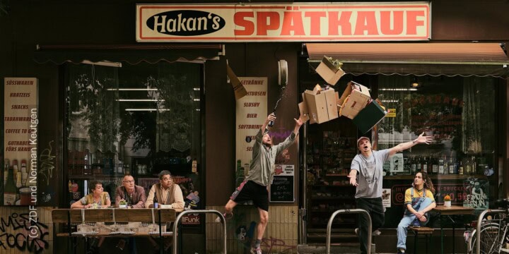 Plakat zur Serie "Späti" für die ZDF Mediathek. Zu sehen sind die Figuren der Serie, die gelangweilt vor "Hakan's Spätkauf" sitzen. Die Hauptfigur, gespielt von Wilson Ochsenknecht, stolpert aus der Tür des Spätis. Zu sehen sind verschiedene Pappkisten, die in der Luft fliegen. Es fallen ein paar Gegenstände aus den Kisten.