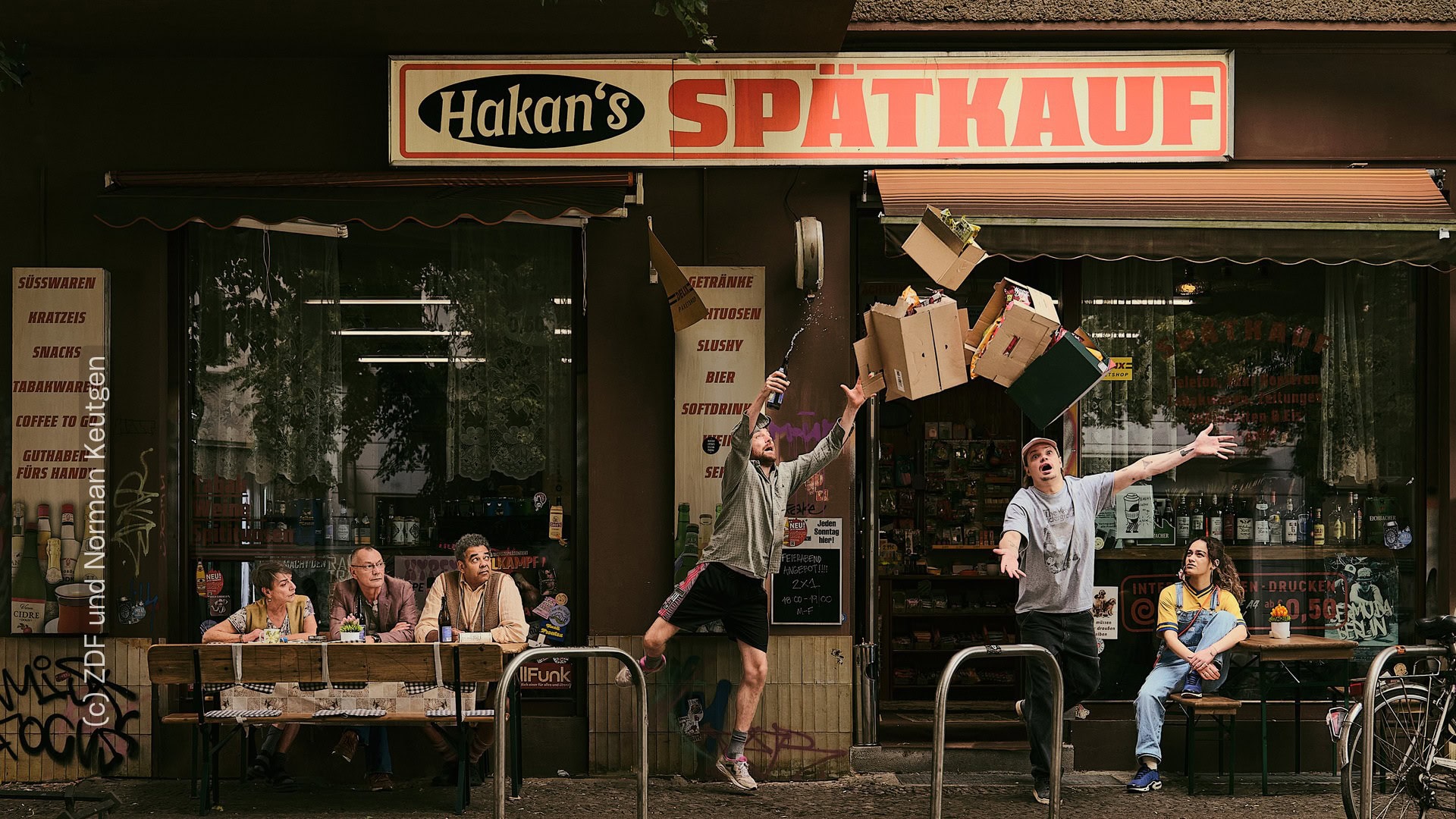 Plakat zur Serie "Späti" für die ZDF Mediathek. Zu sehen sind die Figuren der Serie, die gelangweilt vor "Hakan's Spätkauf" sitzen. Die Hauptfigur, gespielt von Wilson Ochsenknecht, stolpert aus der Tür des Spätis. Zu sehen sind verschiedene Pappkisten, die in der Luft fliegen. Es fallen ein paar Gegenstände aus den Kisten.