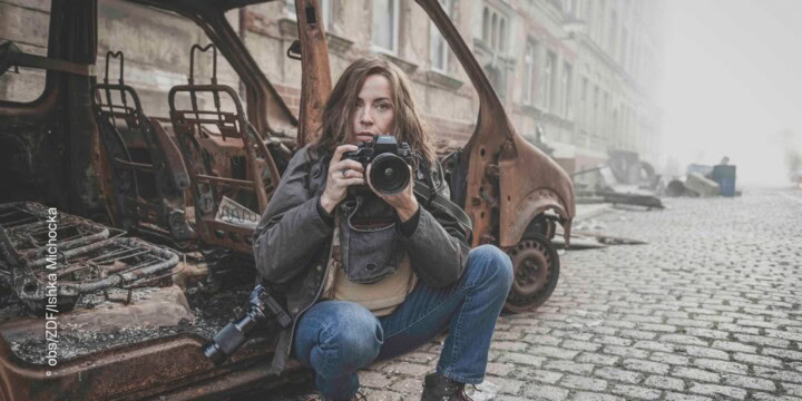 Eine Frau sitzt in der Hocke vor einem ausgebrannten Fahrzeug in einer Stadt. Sie blickt in die Kamera und hält dabei selber eine Spiegelreflexkamera in der Hand.