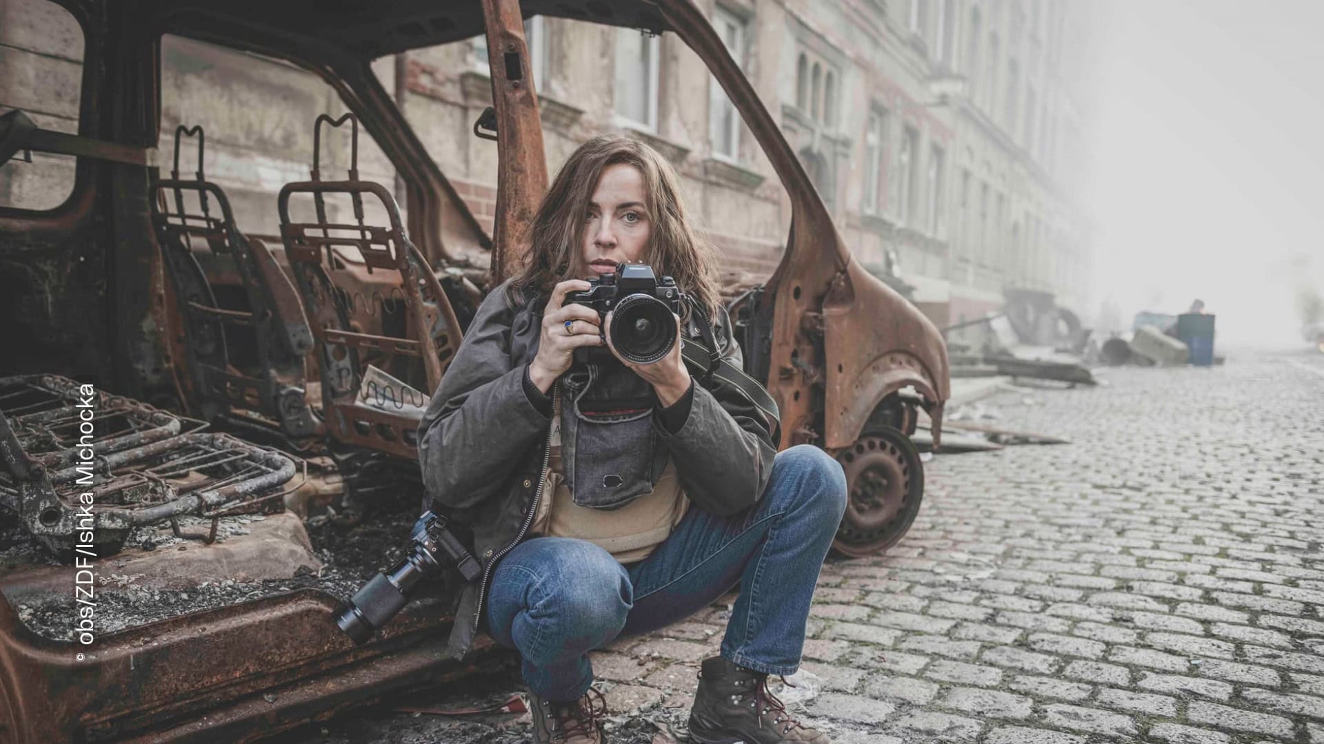 Eine Frau sitzt in der Hocke vor einem ausgebrannten Fahrzeug in einer Stadt. Sie blickt in die Kamera und hält dabei selber eine Spiegelreflexkamera in der Hand.