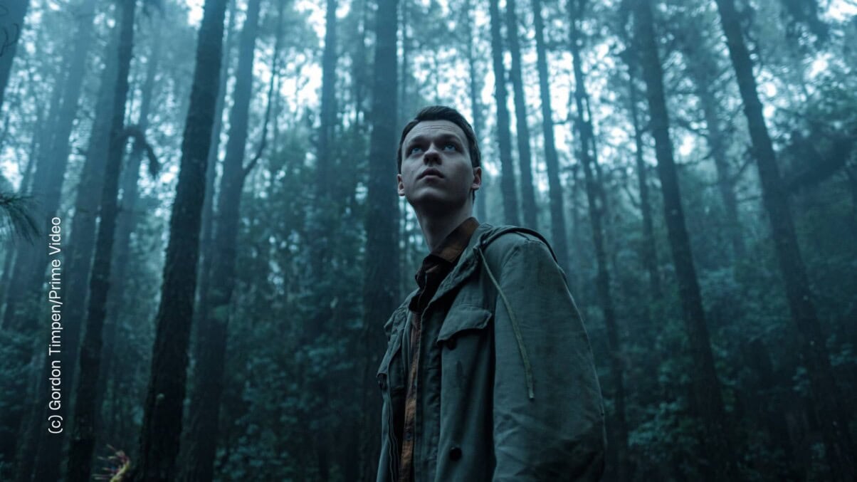 Ein junger Mann steht in einem düsteren Wald und blickt in die Ferne.