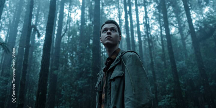 Ein junger Mann steht in einem düsteren Wald und blickt in die Ferne.