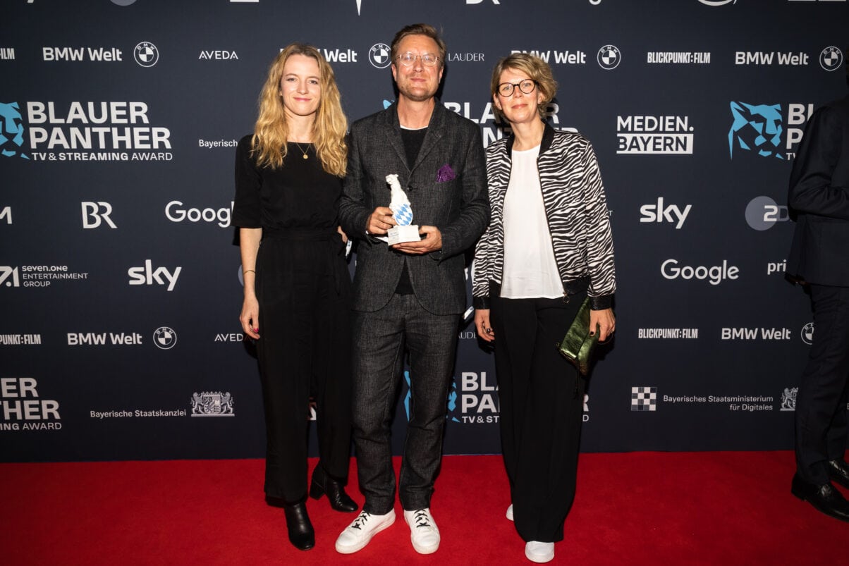 Preisträger Carl Gierstorfer (in der Mitte) mit Mareike Müller (Idee) und Antje Böhmert (Produktion) vor der Pressewand des Blauen Panthers 2022