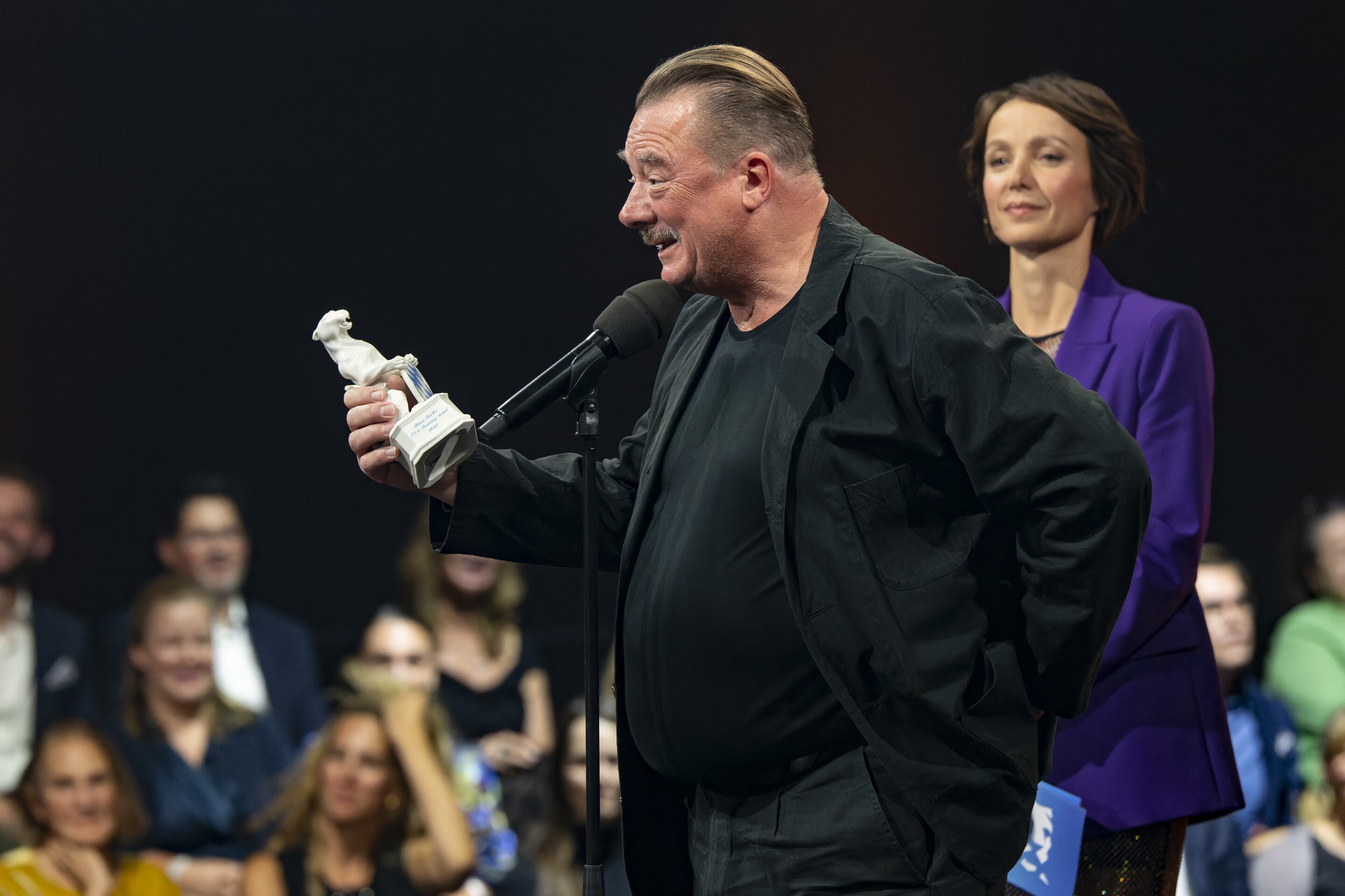 Peter Kurth mit dem Preis in der Hand, vor dem Mikrofon. Er hält gerade seine Dankesrede in der Show 2022.