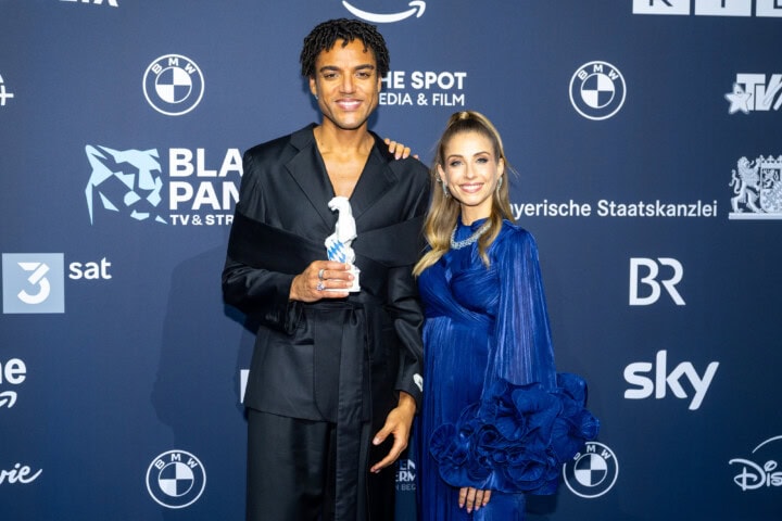 Lamin Leroy Gibba (l.) freut sich mit Moderatorin Cathy Hummels (r.) über den Blauen Panther 2025 in der Kategorie Fiktion als Hauptdarsteller, Creator, Ko-Produzent und Headautor von „Schwarze Früchte“ (Jünglinge Film & Studio Zentral / ARD Degeto Film für die ARD).